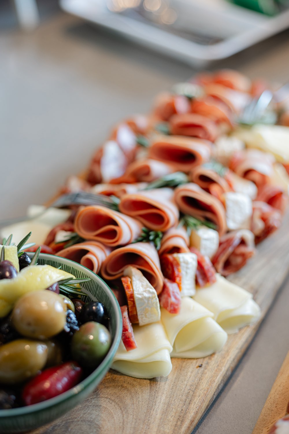 A long wooden charcuterie board of rolled Italian meats, sliced cheeses and brie with fresh rosemary, next to a bowl of mixed olives